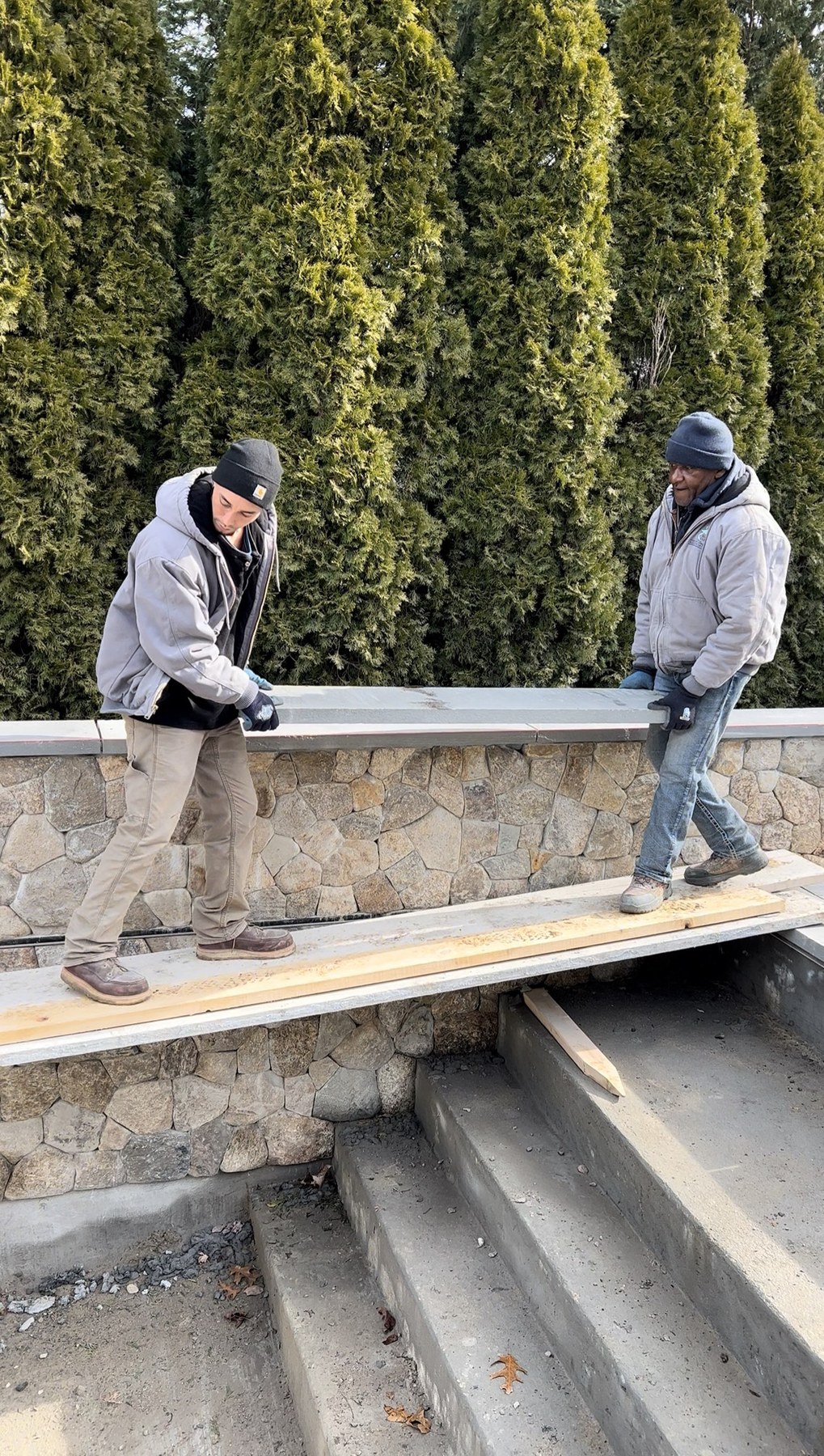 Two Blue Claw Associates crew members carrying a steel beam across a hand-set fieldstone retaining wall on a winter job site
