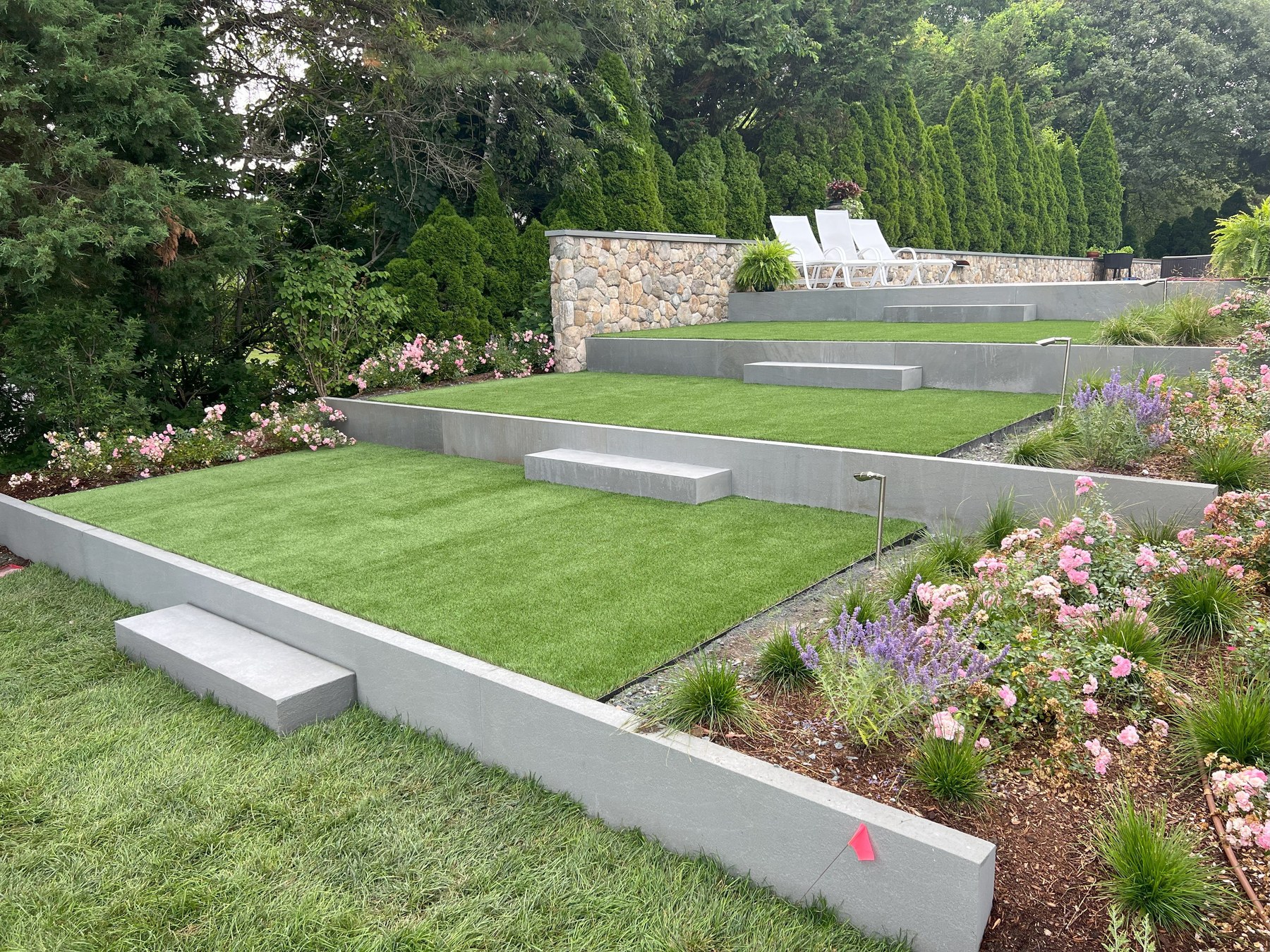 A tiered backyard terrace with cast-concrete walls, perfectly clipped lawns, fieldstone backdrop, and lavender and rose plantings